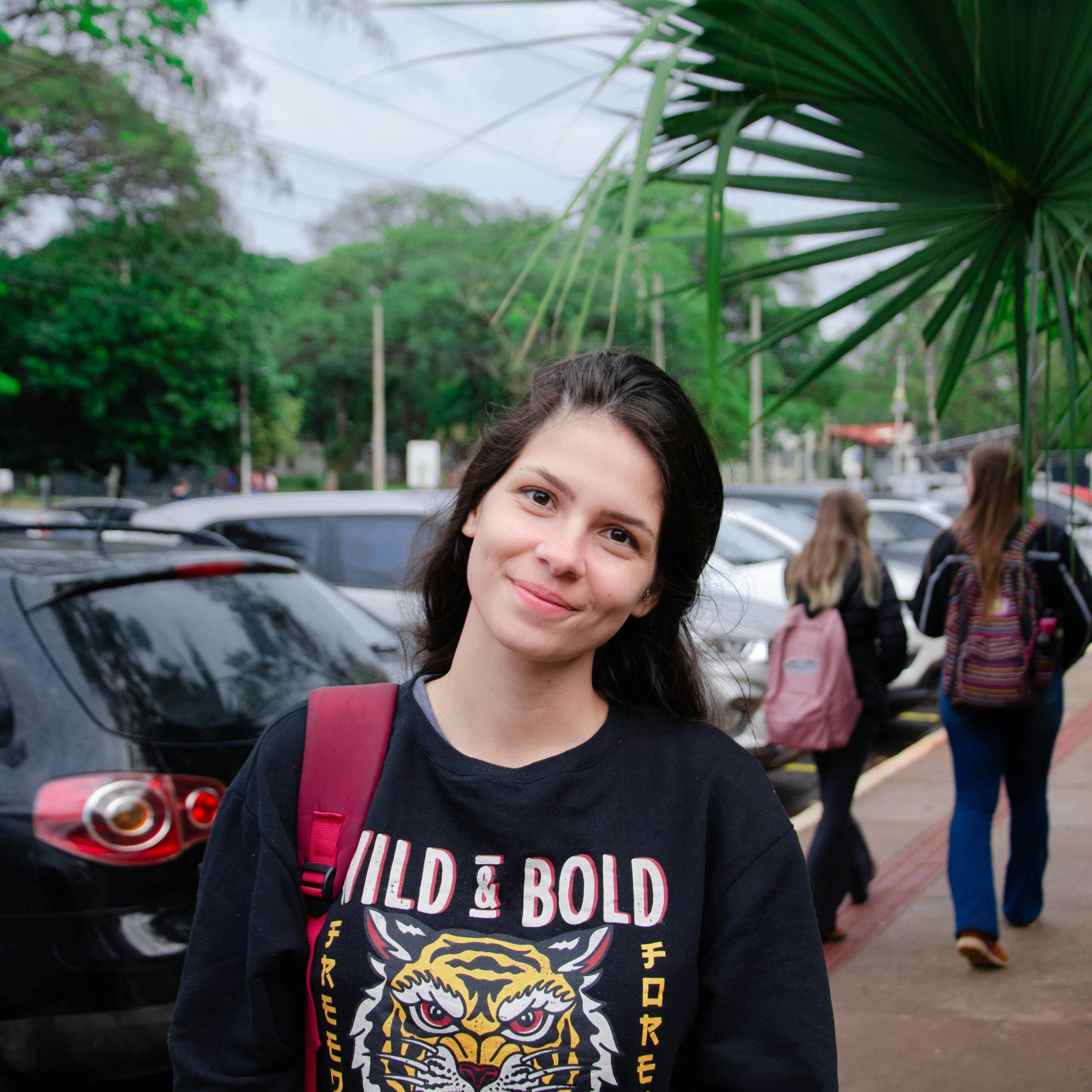 Young woman smiling on a street outdoors, standing near cars and greenery, wearing a casual black sweatshirt.