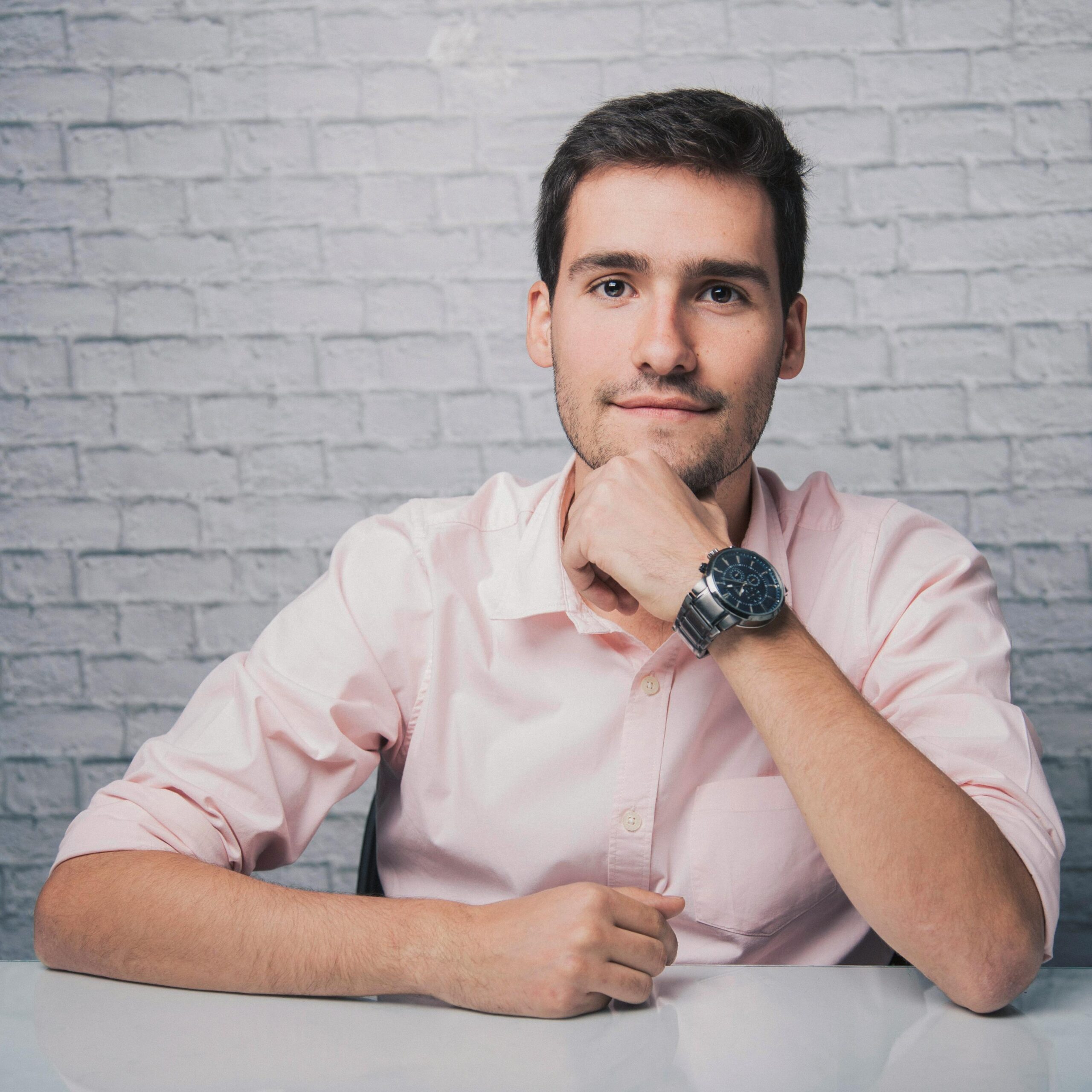 Positive young male in shirt with wristwatch sitting at desk with hand at chin while smiling and looking at camera in room with white brick wall